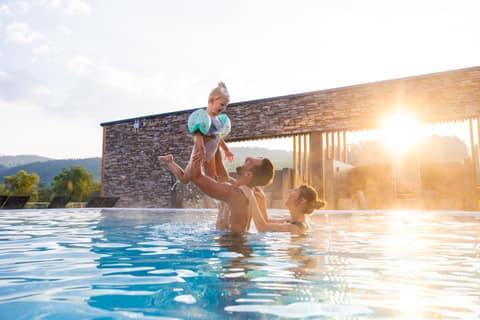 Vater wirft sein Kind im Außenpool in die Luft, im Hintergrund die Abendsonne