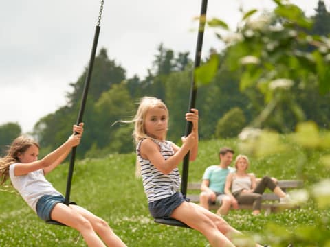 Zwei Mädchen schaukeln auf der Wiese, die Eltern sitzen im Hintergrund auf einer Bank